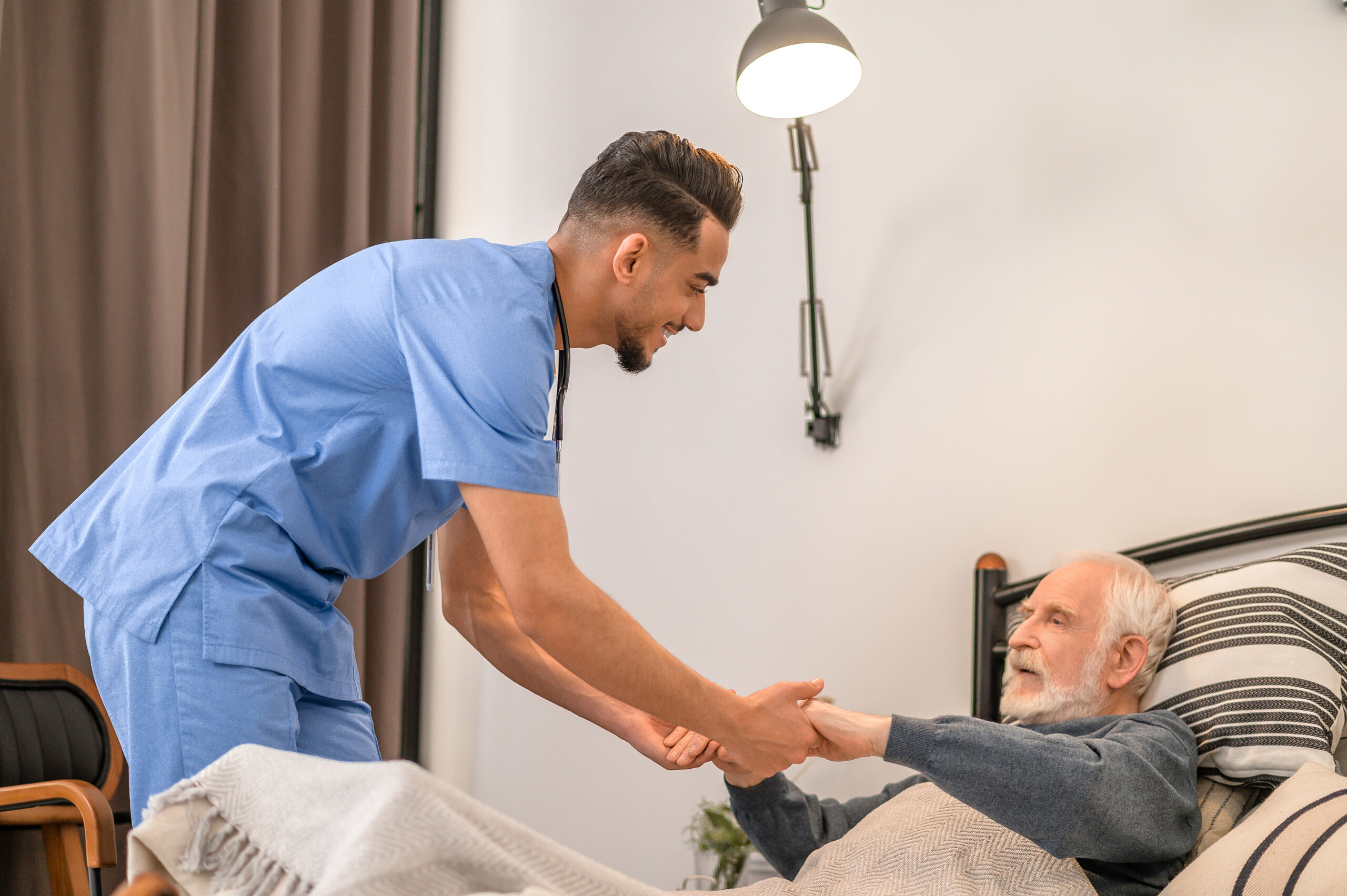 Experienced in-home male nurse in uniform helping an aged patient to rise from the bed